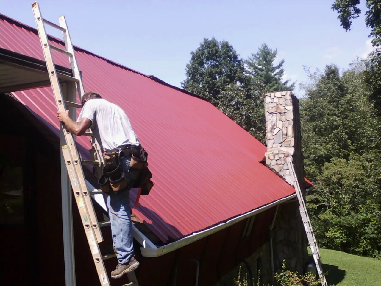 Roofer on a ladder beside a red metal roof and stone chimney — Soffit & Fascia Repair by Roofing Brothers in Mifflintown